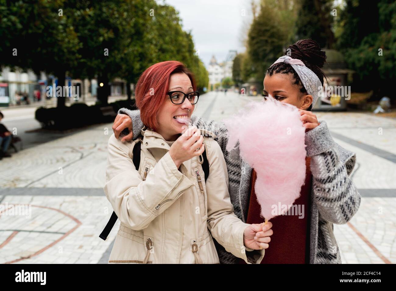 Stylish cheerful multiethnic girl friends walking embracing and eating pink candy cotton in street Stock Photo