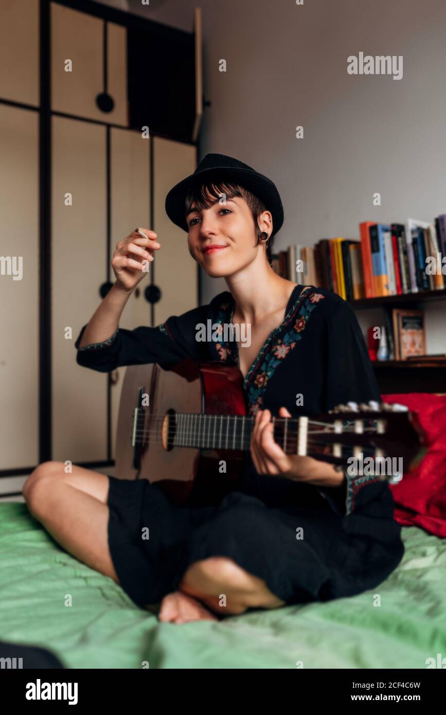 Relaxed Woman in stylish clothes and hat sitting on bed with crossed ...