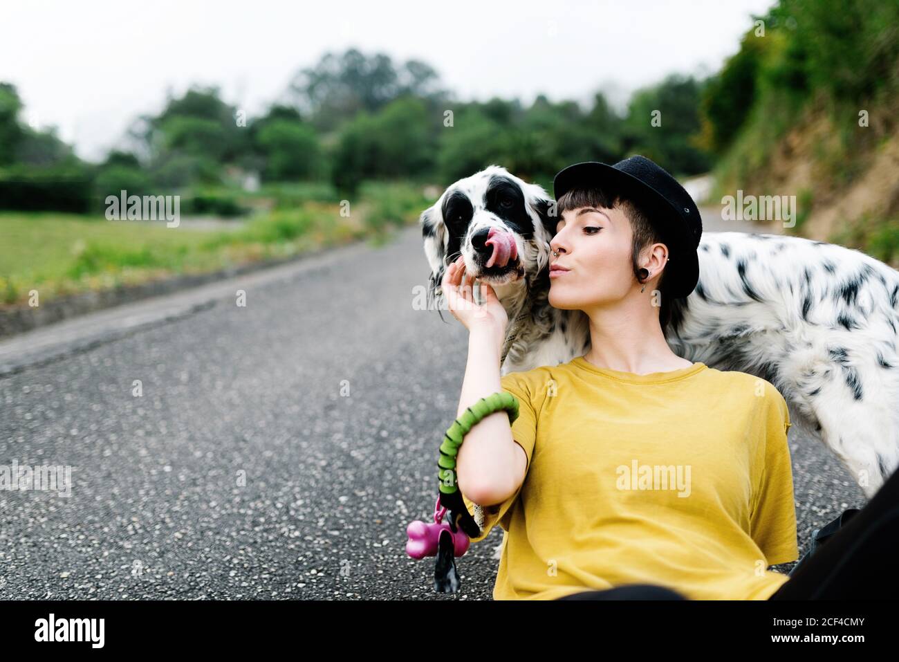 Positive young lady in casual clothes and black hat resting on ground with dog during walk in park Stock Photo