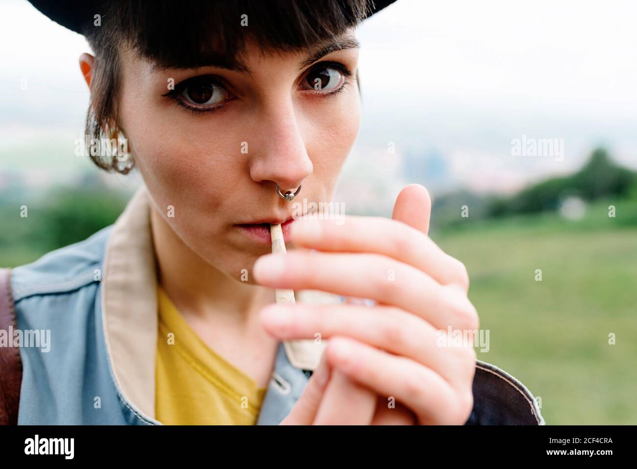 Crop informal lady in casual clothes with piercing in nose lighting cigarette while standing on street and looking at camera Stock Photo