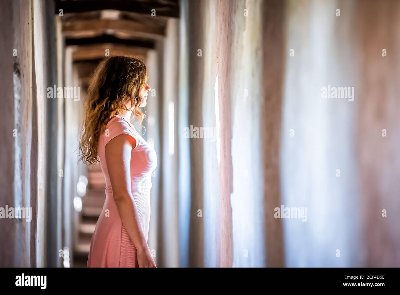 Young woman in pink dress side profile standing looking through window of wall of fortress tunnel passage in Castiglione del Lago in Italy Stock Photo