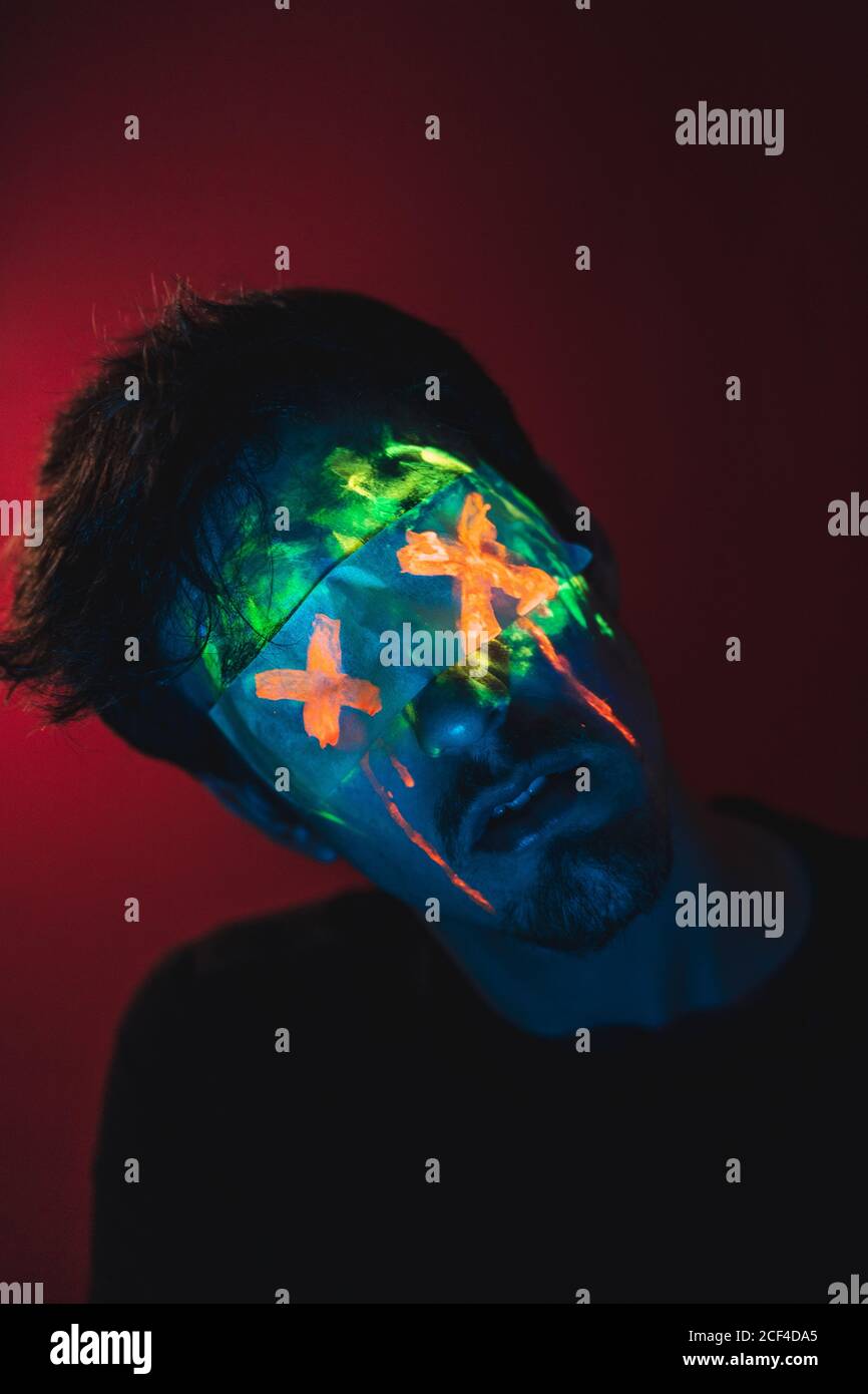 man with blue hair and green radiant face wearing tape with painted X on red background Stock Photo