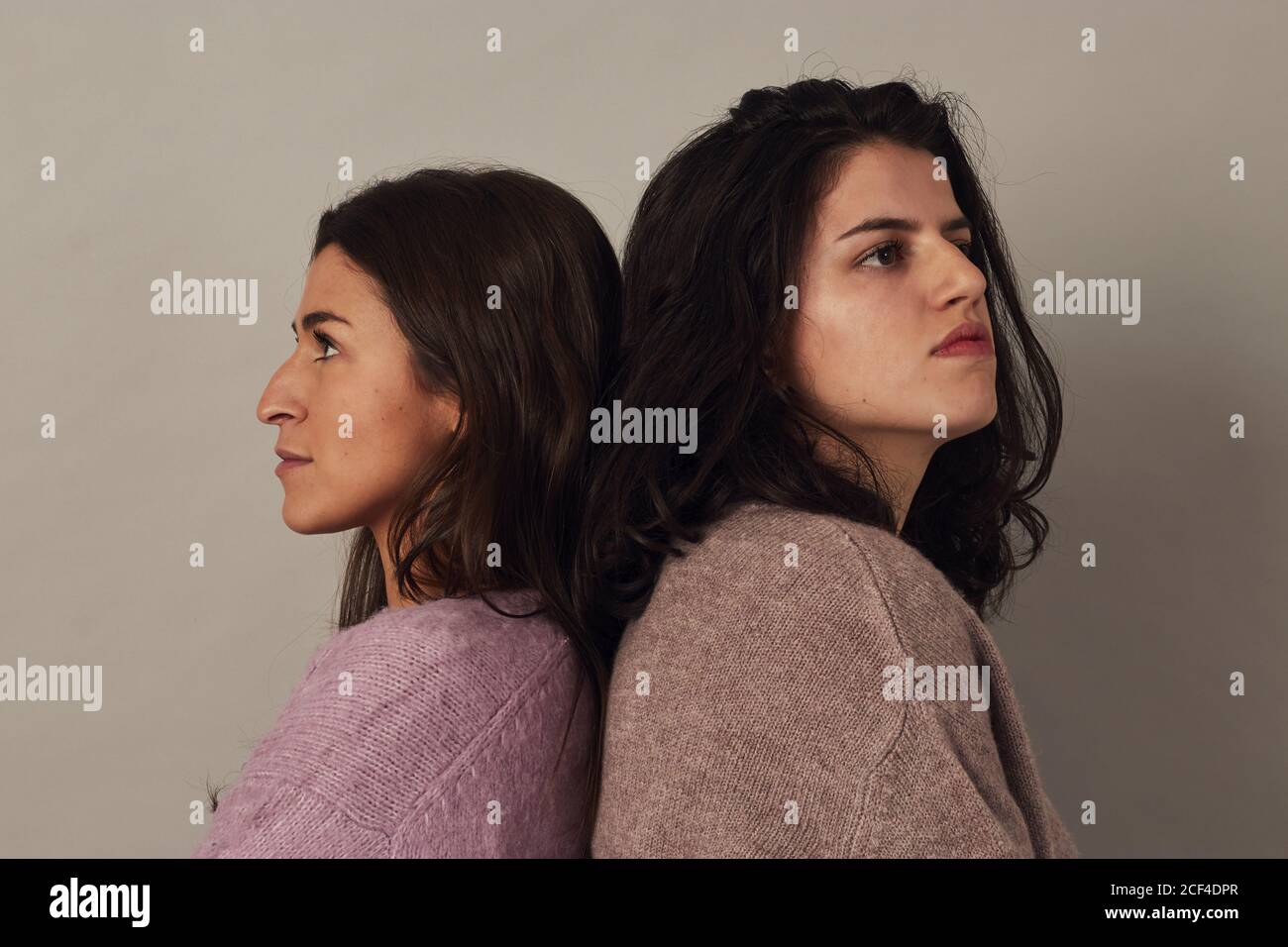 Side view of satisfied young charming dark haired women wearing sweaters and looking at camera while standing together on gray background in studio Stock Photo