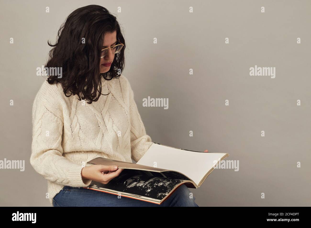 Serious youthful dark haired woman in glasses and white sweater looking through book while sitting in studio Stock Photo