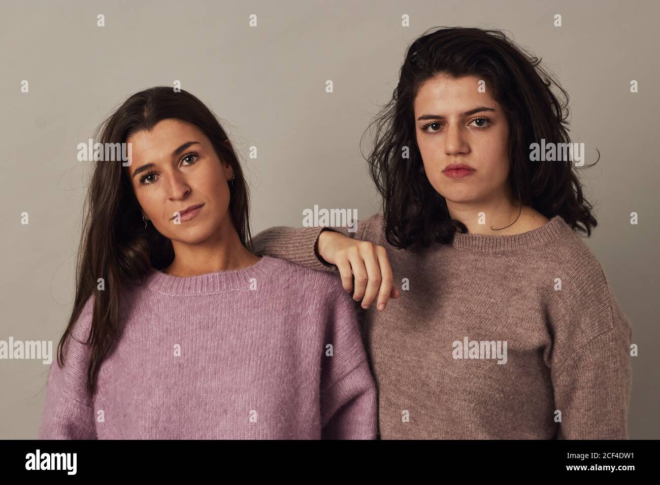 Satisfied young charming dark haired women wearing sweaters and looking at camera while standing together on gray background in studio Stock Photo