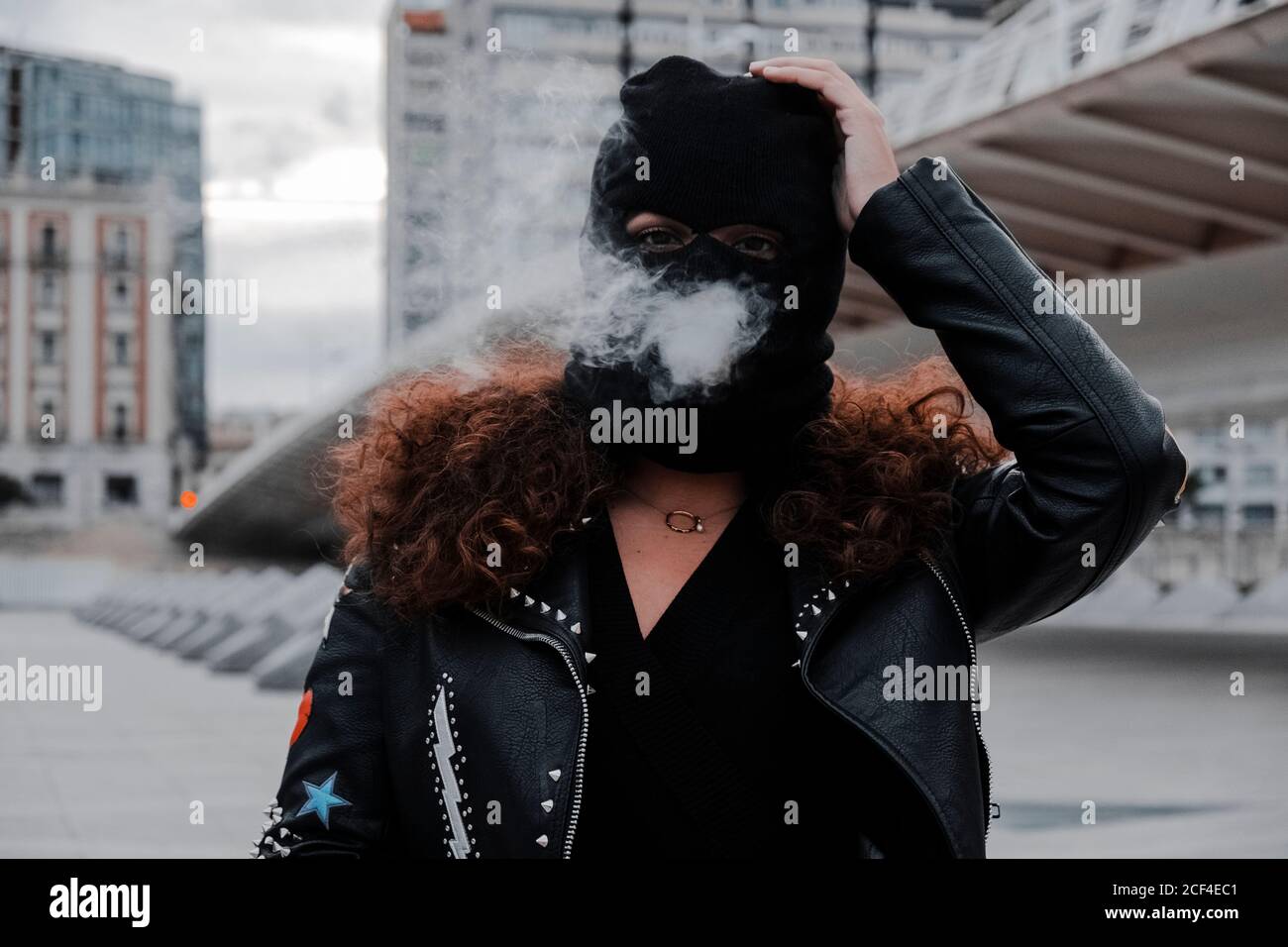 Woman in balaclava and rocker styled black leather jacket with stripes on the middle of the street smoking Stock Photo