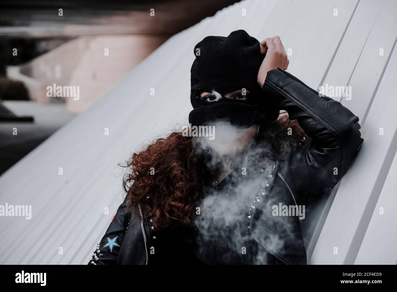 Woman in balaclava and rocker styled black leather jacket with stripes  leaning back on concrete wall and smoking Stock Photo - Alamy