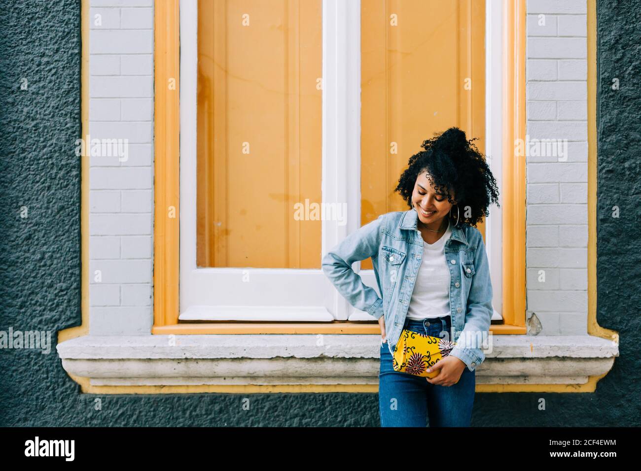 Trendy ethnic Woman in jeans and denim jacket holding fashion handbag and leaning on bright windowsill outdoors Stock Photo