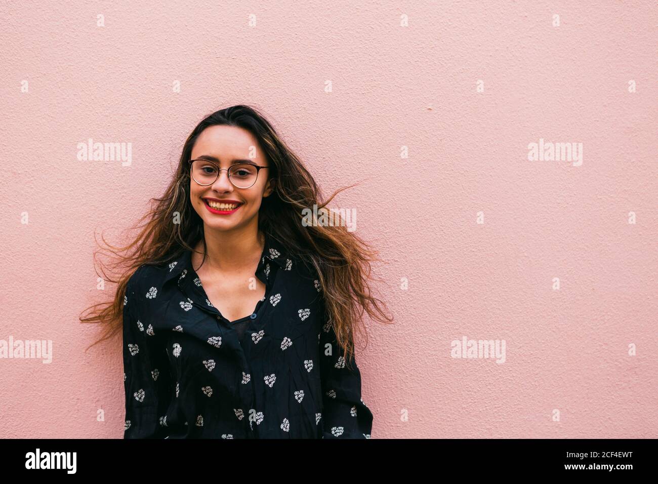 Charming teen Woman in eyeglasses and blue shirt smiling happily at camera standing against pink wall Stock Photo