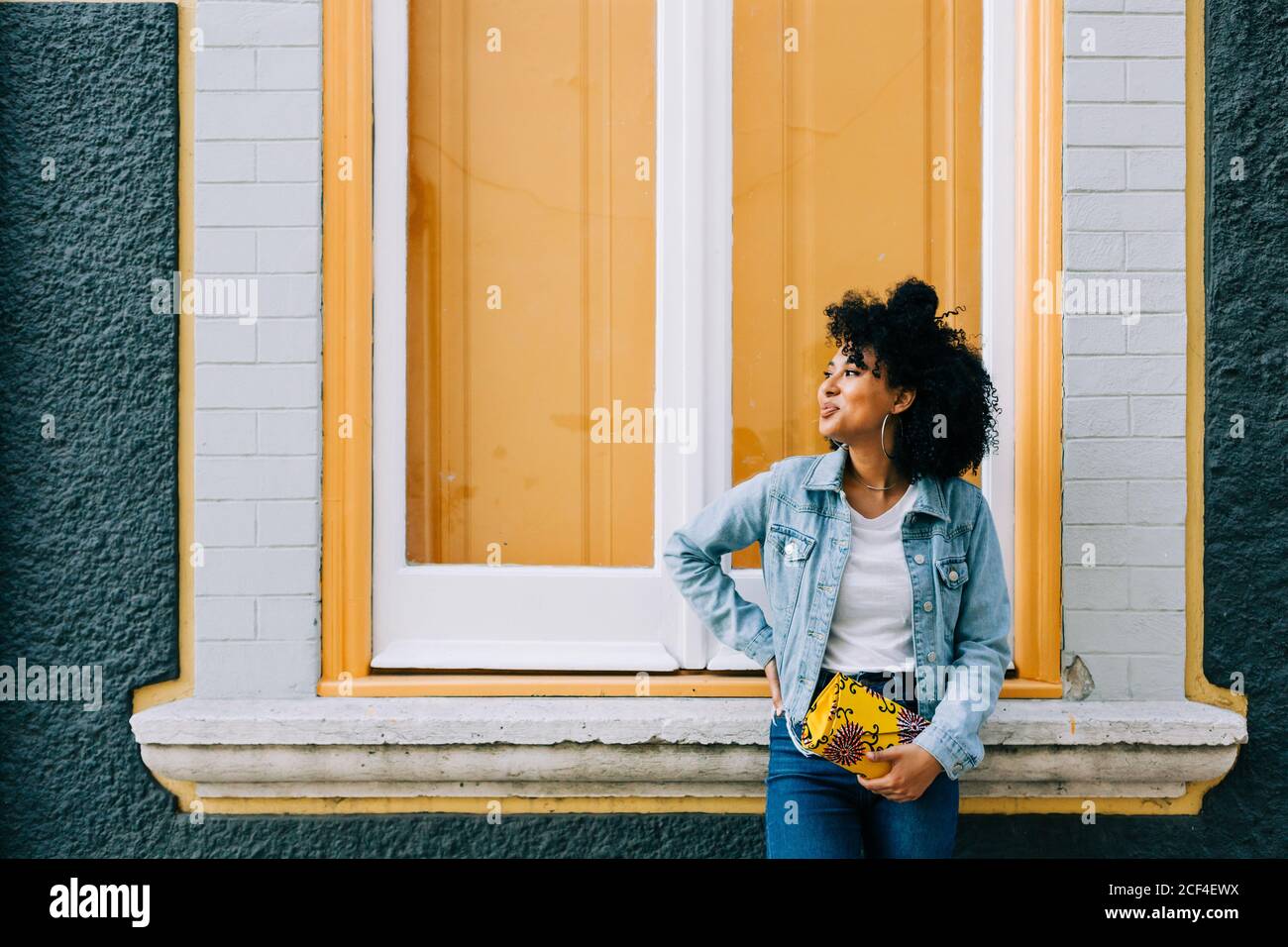 Trendy ethnic Woman in jeans and denim jacket holding fashion handbag and leaning on bright windowsill outdoors Stock Photo