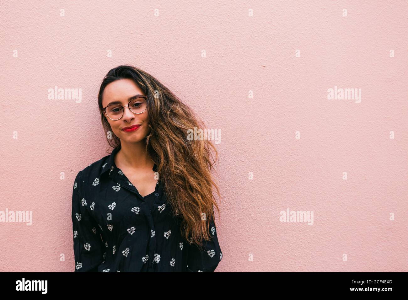 Charming teen Woman in eyeglasses and blue shirt smiling happily at camera standing against pink wall Stock Photo