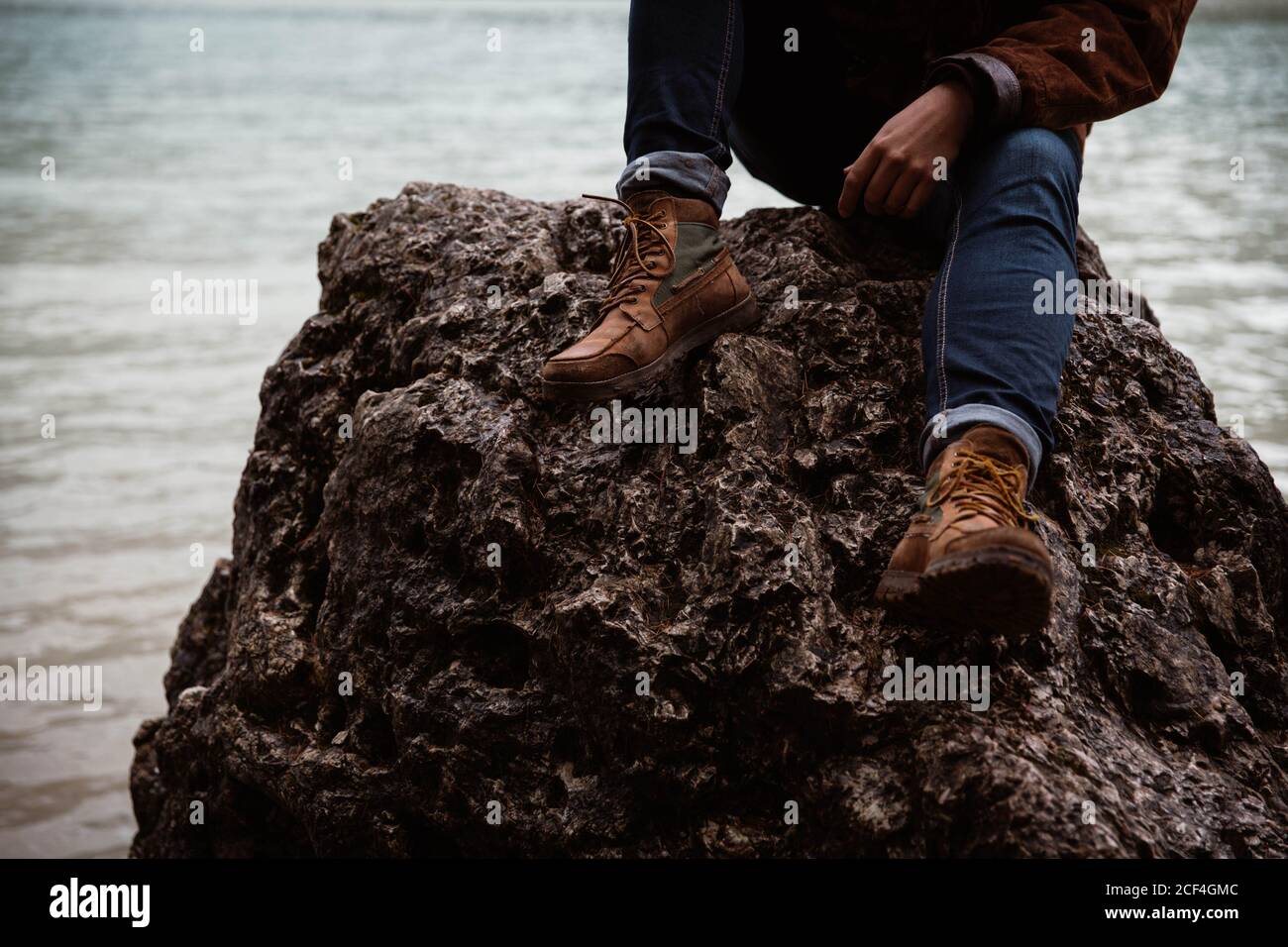 Anonymous cropped man traveler in casual wear sitting in a rock delighting in view of turquoise lake with foggy Dolomites mountains on background at Italy Stock Photo