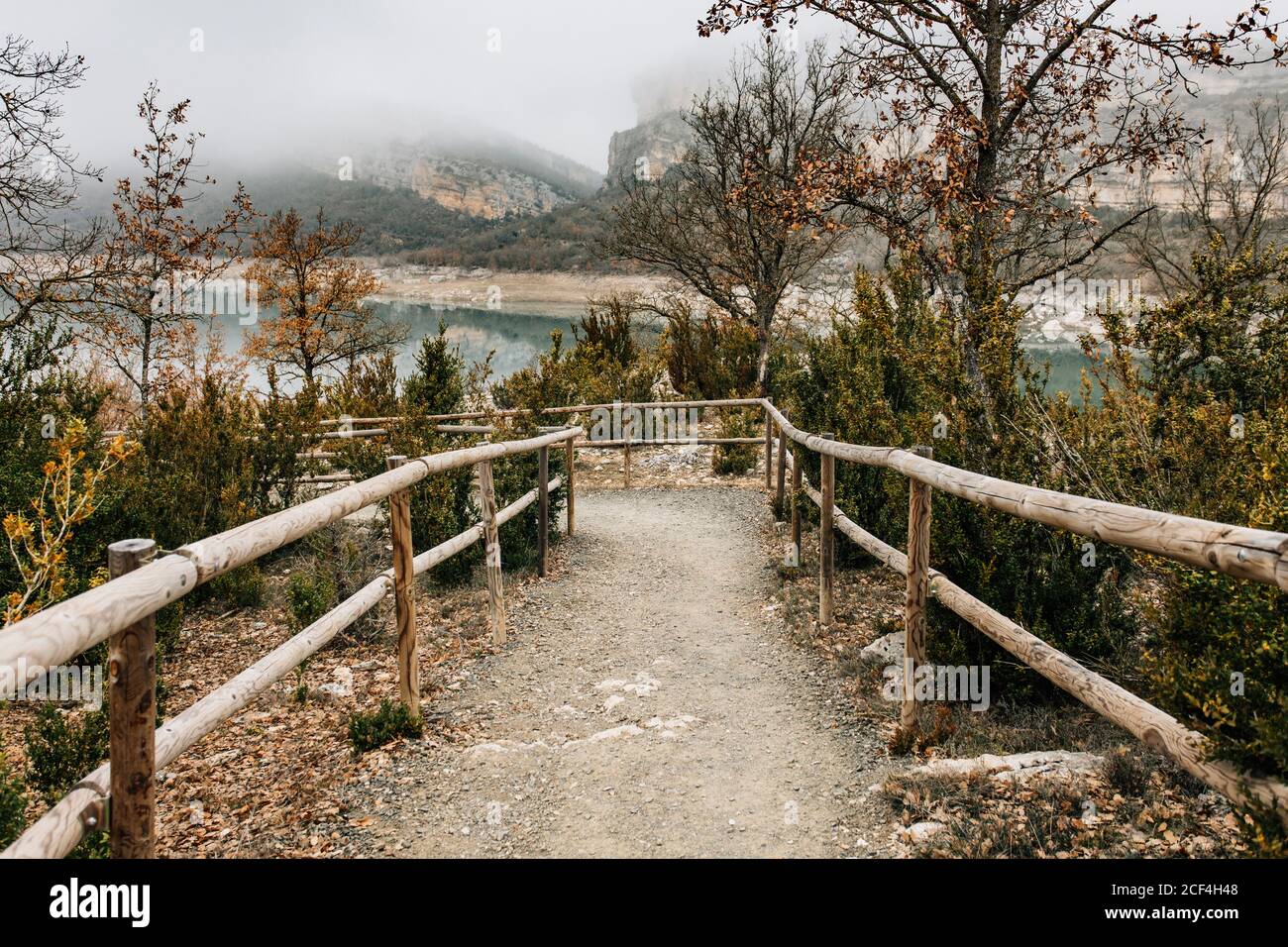 Empty curved path with wooden railing leading among trees with dry foliage on hill slope near mountain lake in foggy day in Montsec Range in Spain Stock Photo