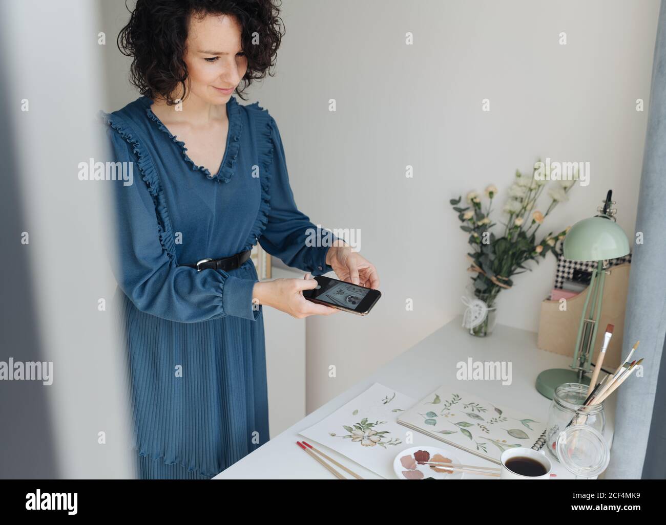 Woman artist in blue dress taking pictures on mobile phone of watercolor works on table in studio Stock Photo