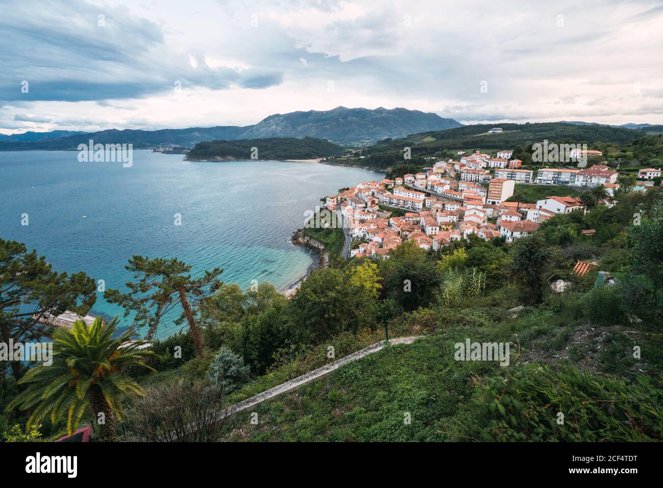 Drone view of peaceful seashore with green trees and small town with red roofs locating near blue sea and mountain ridge against blue cloudy sky Stock Photo
