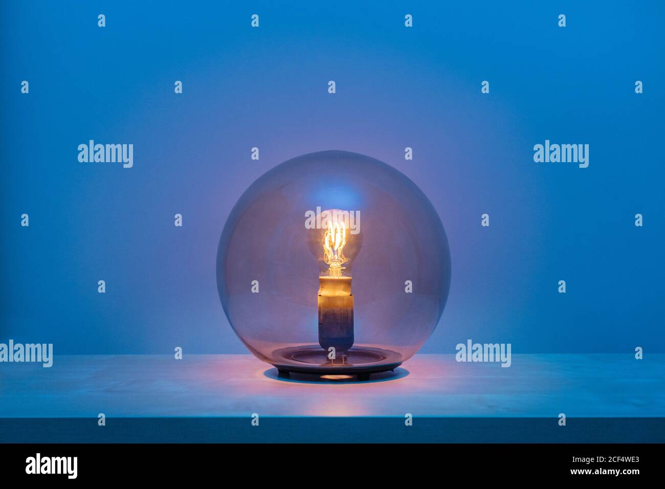 Modern turned on luminaire with light bulb inside thin transparent glass sphere in middle of shelf in blue room at dusk Stock Photo
