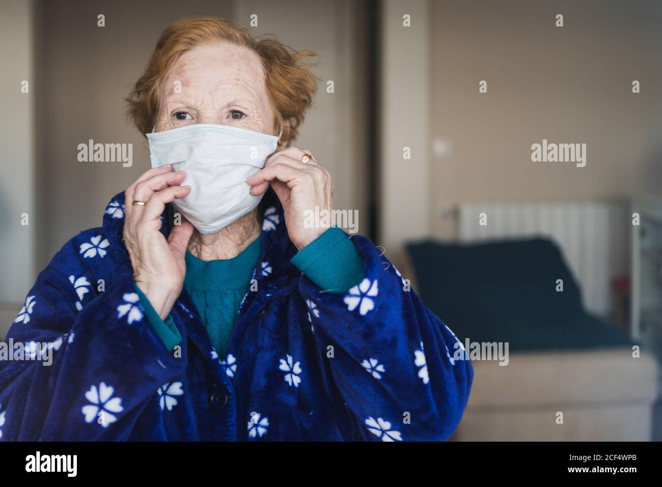 Senior female with red hair in blue robe and medical mask looking at camera while standing in hospital room Stock Photo