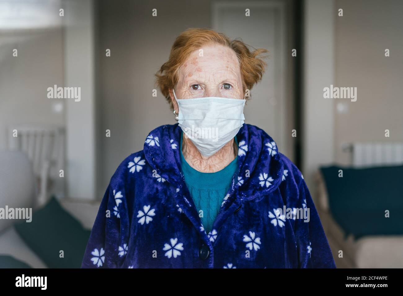 Senior female with red hair in blue robe and medical mask looking at camera while standing in hospital room Stock Photo
