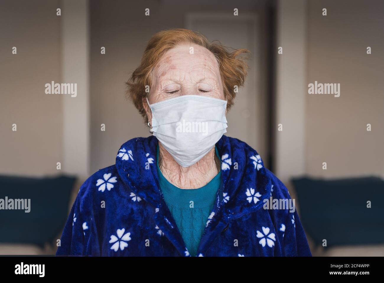 Senior female with red hair in blue robe and medical mask looking at camera while standing in hospital room Stock Photo