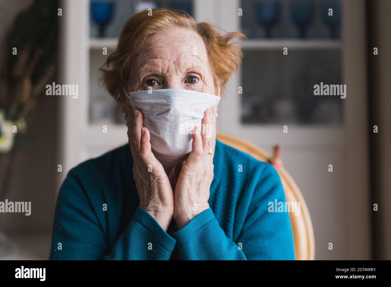 Senior female with red hair in blue robe and medical mask looking at camera while standing in hospital room Stock Photo