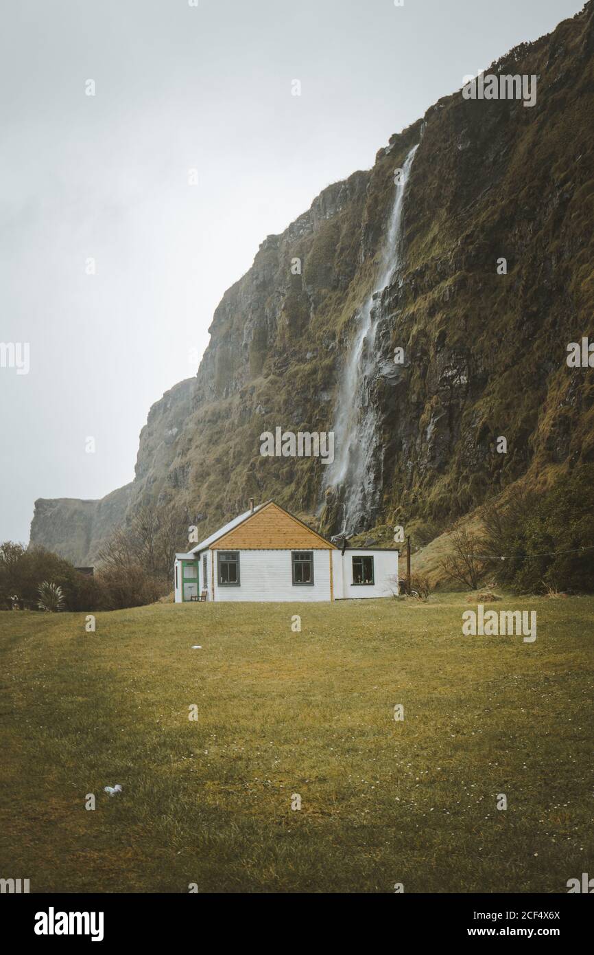 Small country wooden house with white walls and yellow gabled roof located on green meadow at foot of cliff with waterfall against grey cloudy sky in spring day in Northern Ireland Stock Photo