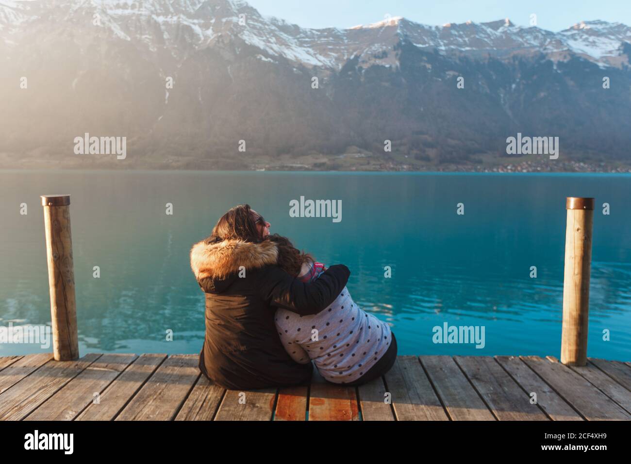 Back view of embracing women sitting on wood pier above turquoise lake in snowy mountains of Switzerland Stock Photo
