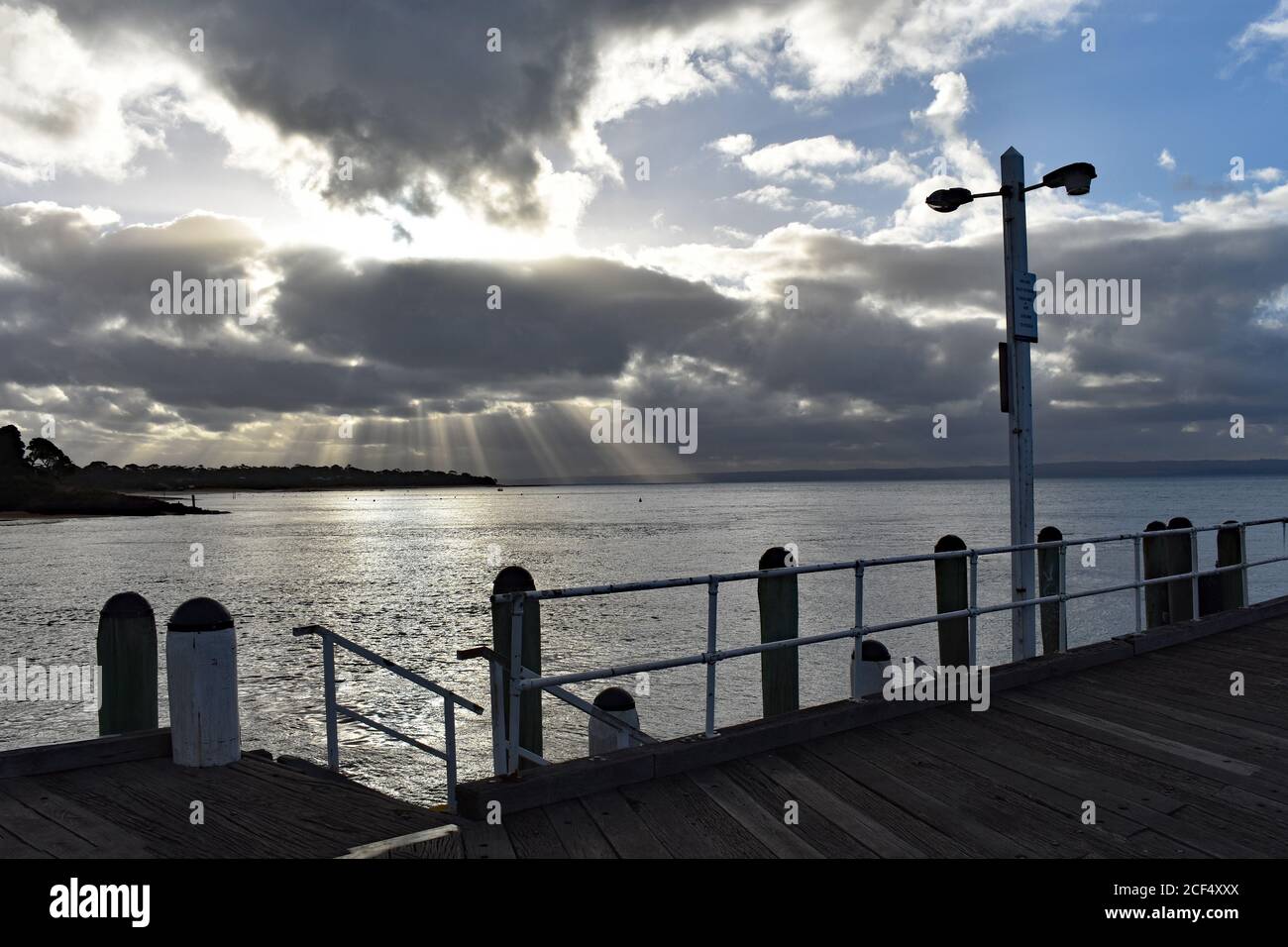Cowes Pier on Phillip Island, Australia.  The sun's rays are shinning through the clouds in the distance and light up the sea surrounding the pier. Stock Photo
