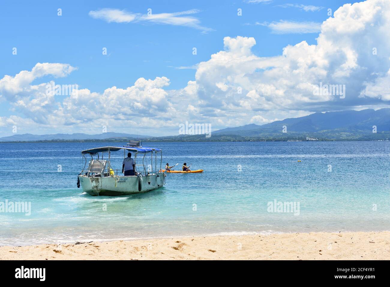 Two Kayakers pass by a small boat  in the waters around South Sea Island, part of the Mamanuca Islands in Fiji.   Golden sands and blue calm sea. Stock Photo