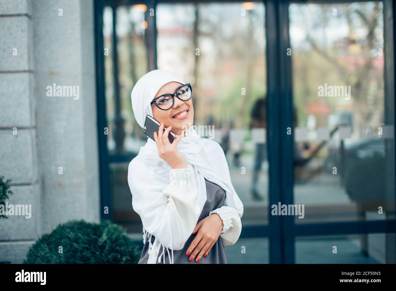 Young woman with glasses wearing white hijab head scarf in city talking ...