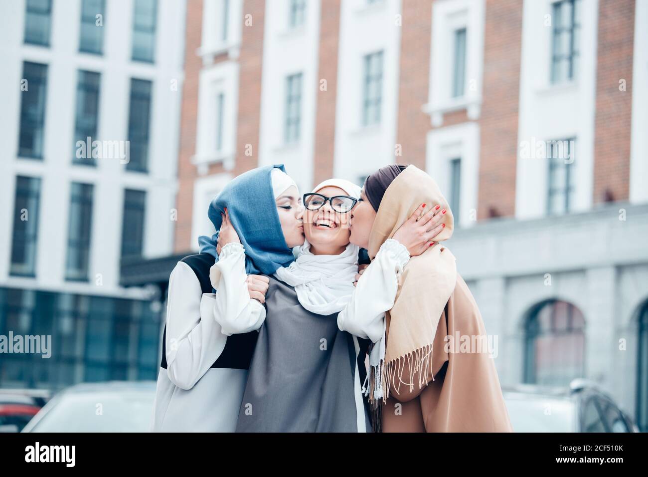 Two funny affectionate muslim women friends laughing and kissing girl in hijab an with glasses outdoors. Stock Photo