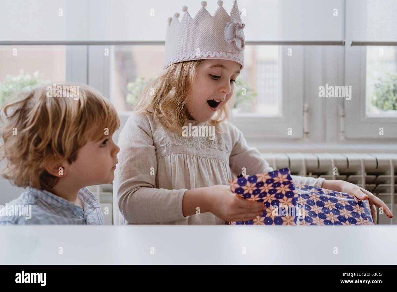 Excited charming girl in handmade crown unwrapping gift box while having birthday celebration at home Stock Photo