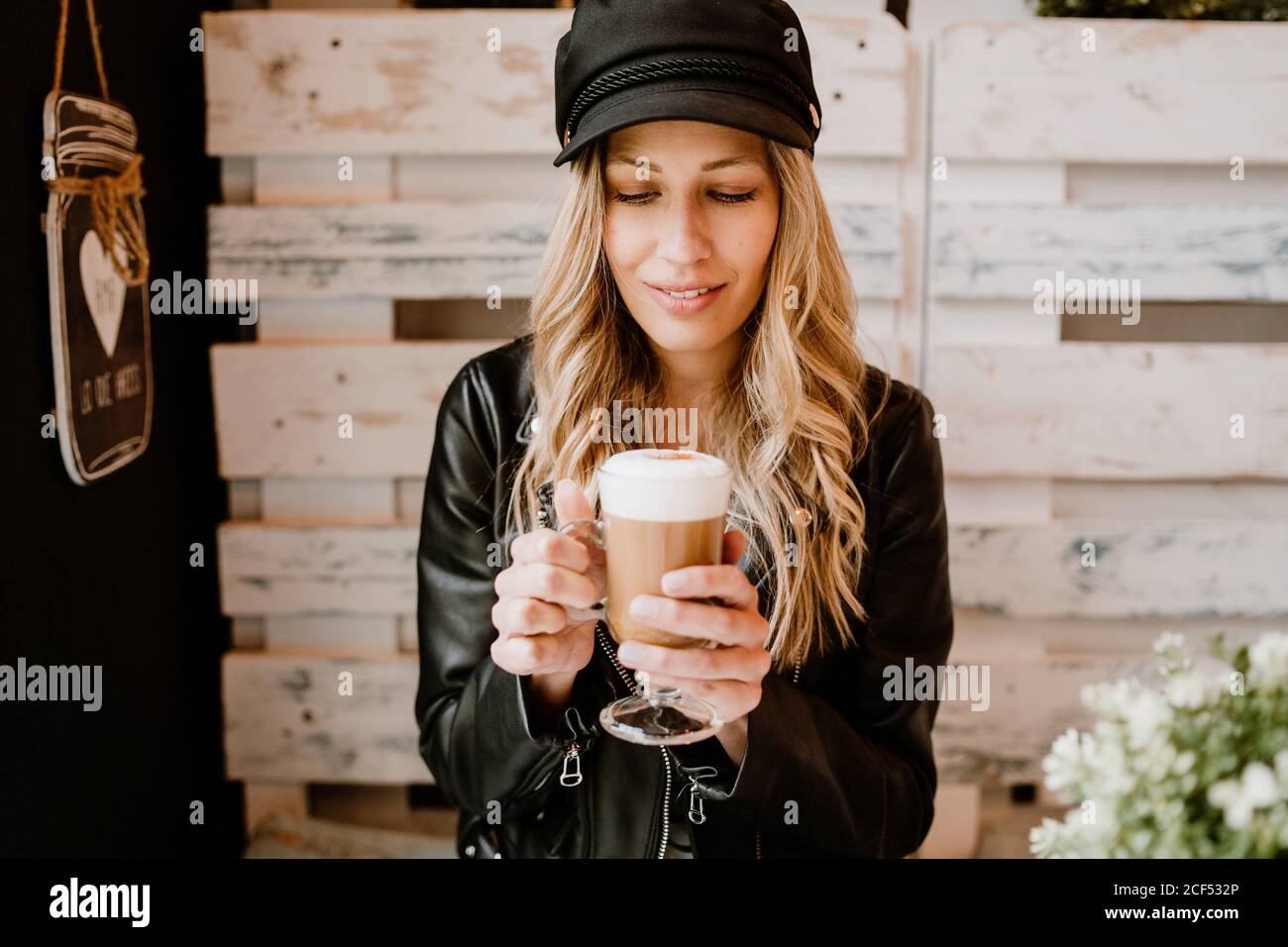 Long haired trendy Woman holding glass of delicious foamy coffee licking lips anticipating pleasure of taste in cafe Stock Photo
