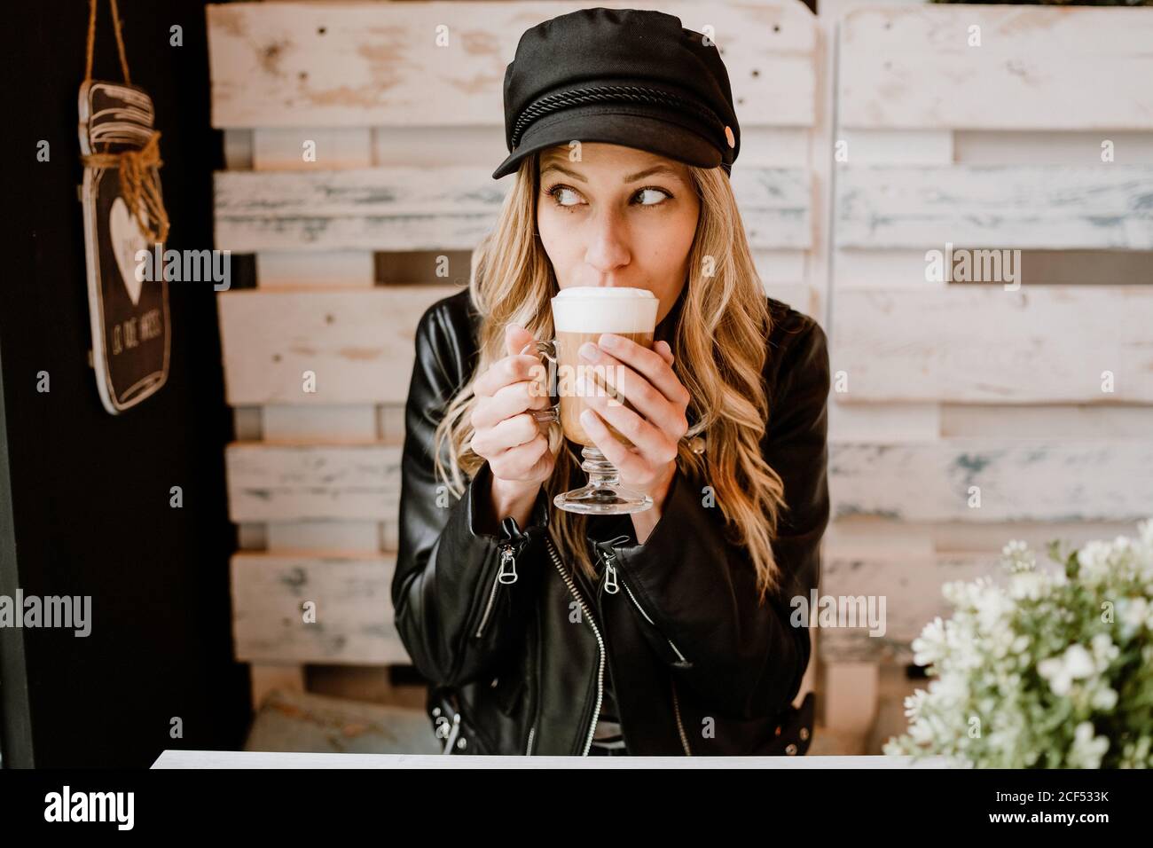 Long haired trendy Woman holding glass of delicious foamy coffee licking lips anticipating pleasure of taste in cafe Stock Photo