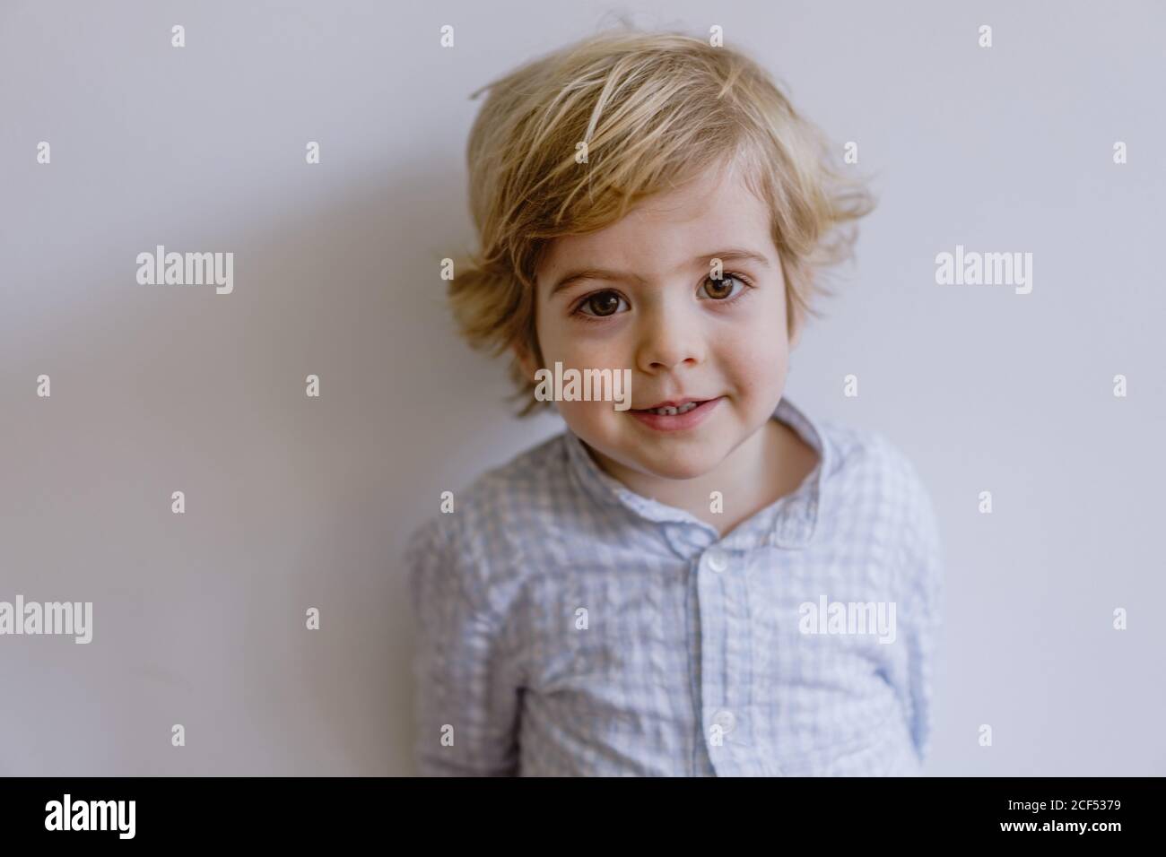 Adorable content child in casual shirt standing near while wall and smiling while looking at camera Stock Photo
