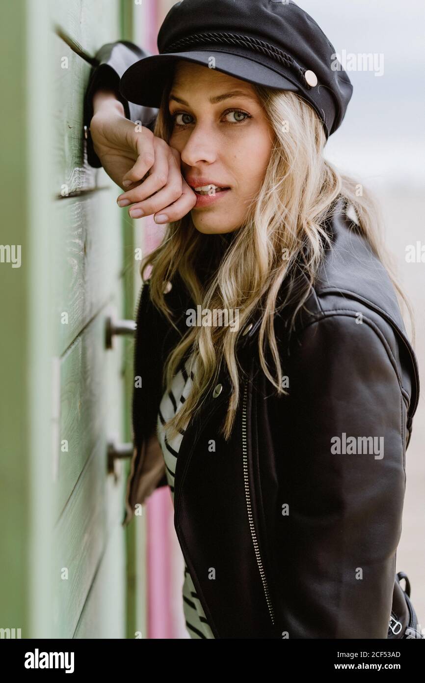 Trendy long haired blonde Woman in black cap and leather jacket smiling  brightly at camera and leaning on wall of wooden beach cabins Stock Photo -  Alamy