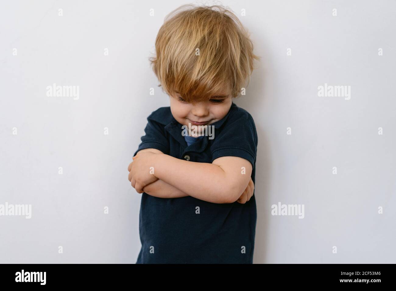 Annoyed blond little boy in casual clothes looking down with dissatisfaction while standing on white wall with crossed arms Stock Photo