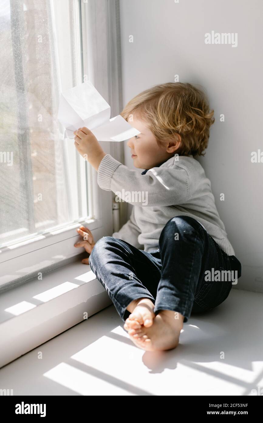 Side view of blonde little child in casual clothes playing with paper airplane while sitting barefoot on window sill on sunny day Stock Photo