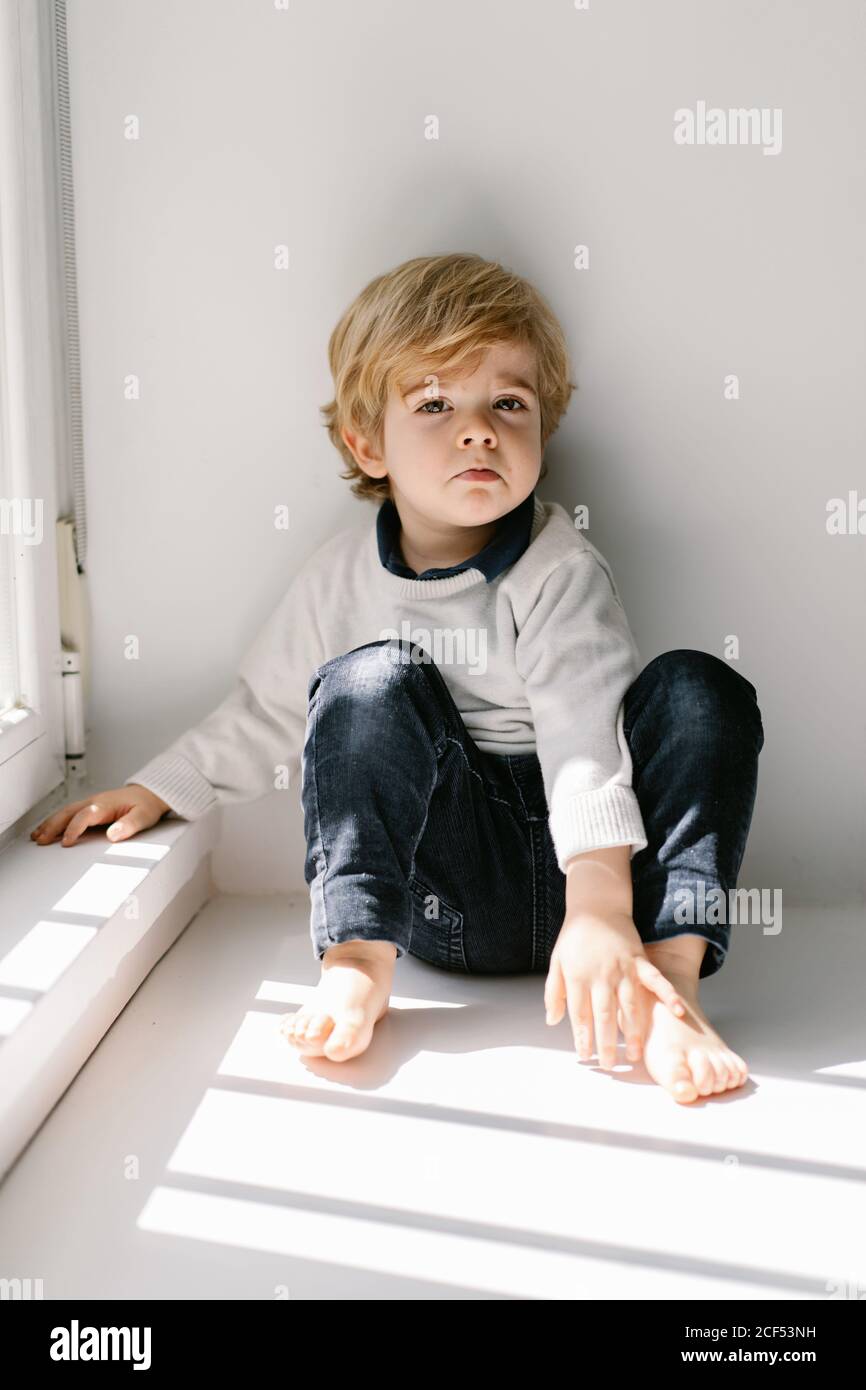 Annoyed blond little boy looking at camera with dissatisfaction while sitting on sill leaning on white wall Stock Photo