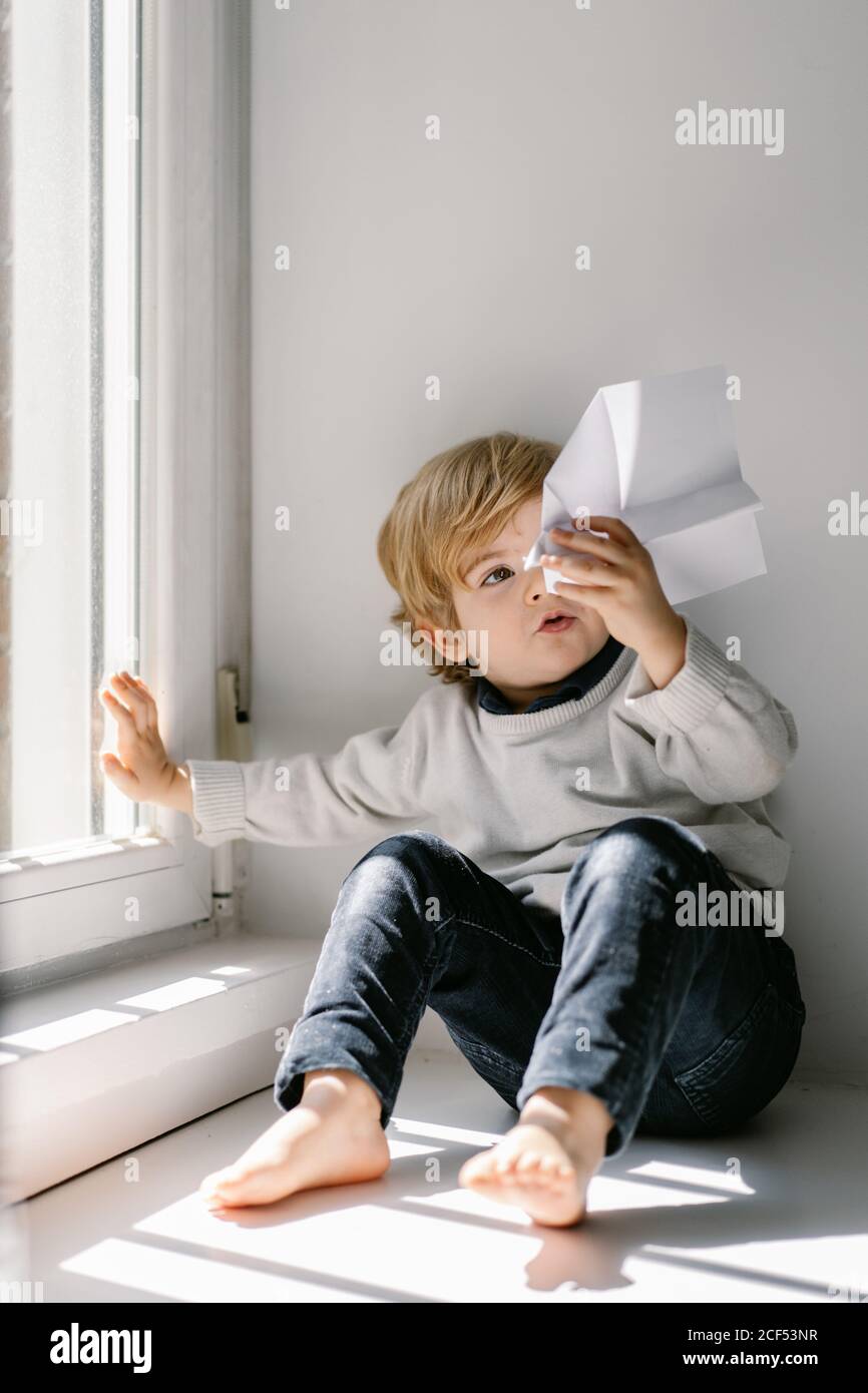 Happy blonde little child in casual clothes playing with paper airplane while sitting barefoot on window sill on sunny day Stock Photo