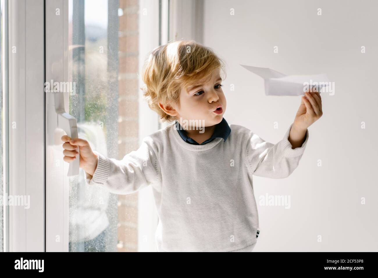 Happy blond child in casual clothes playing with paper airplane while sitting barefoot on window sill on sunny day Stock Photo