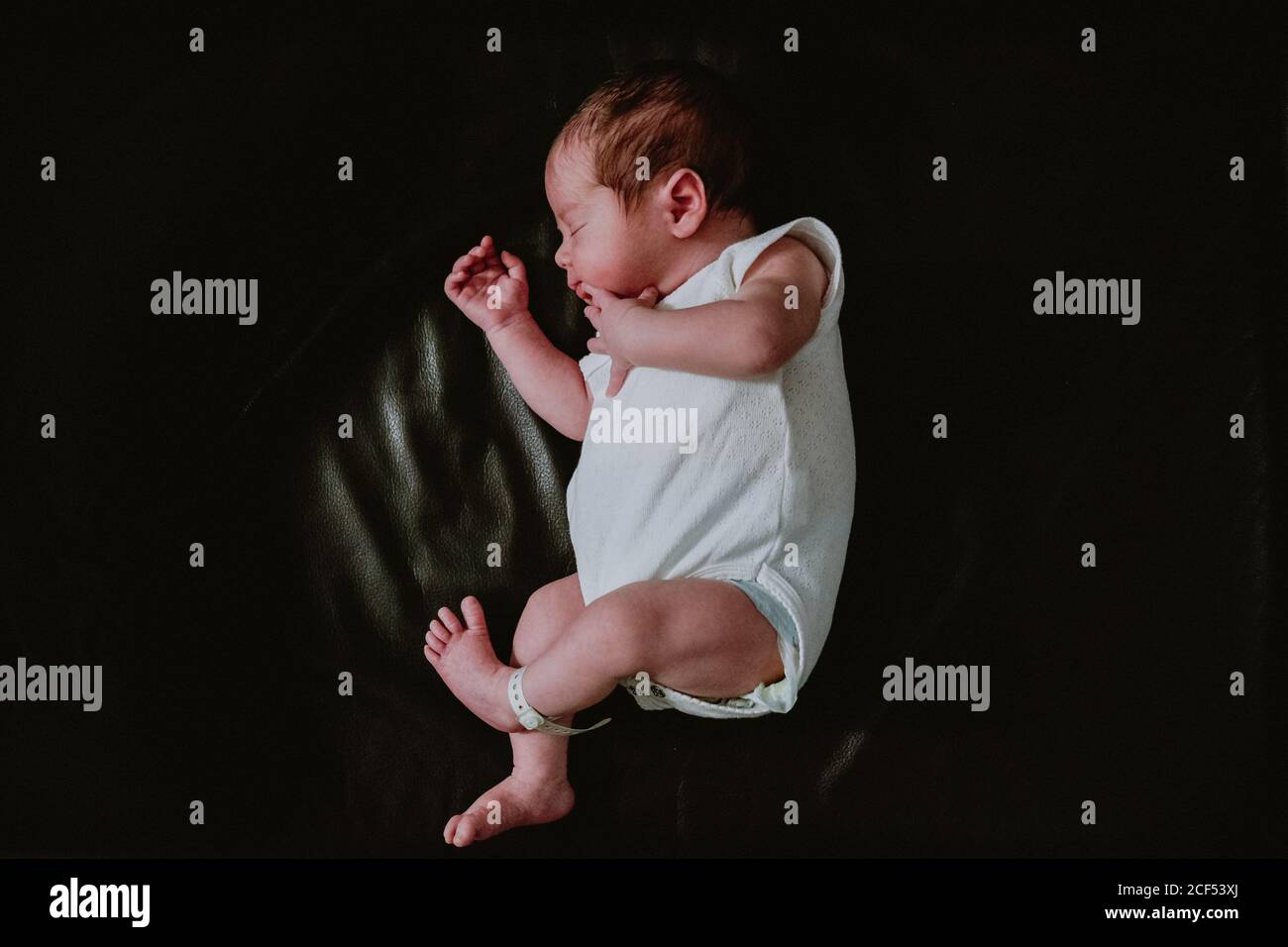 From above of cute tiny newborn baby in white bodysuit sleeping peacefully on black background Stock Photo