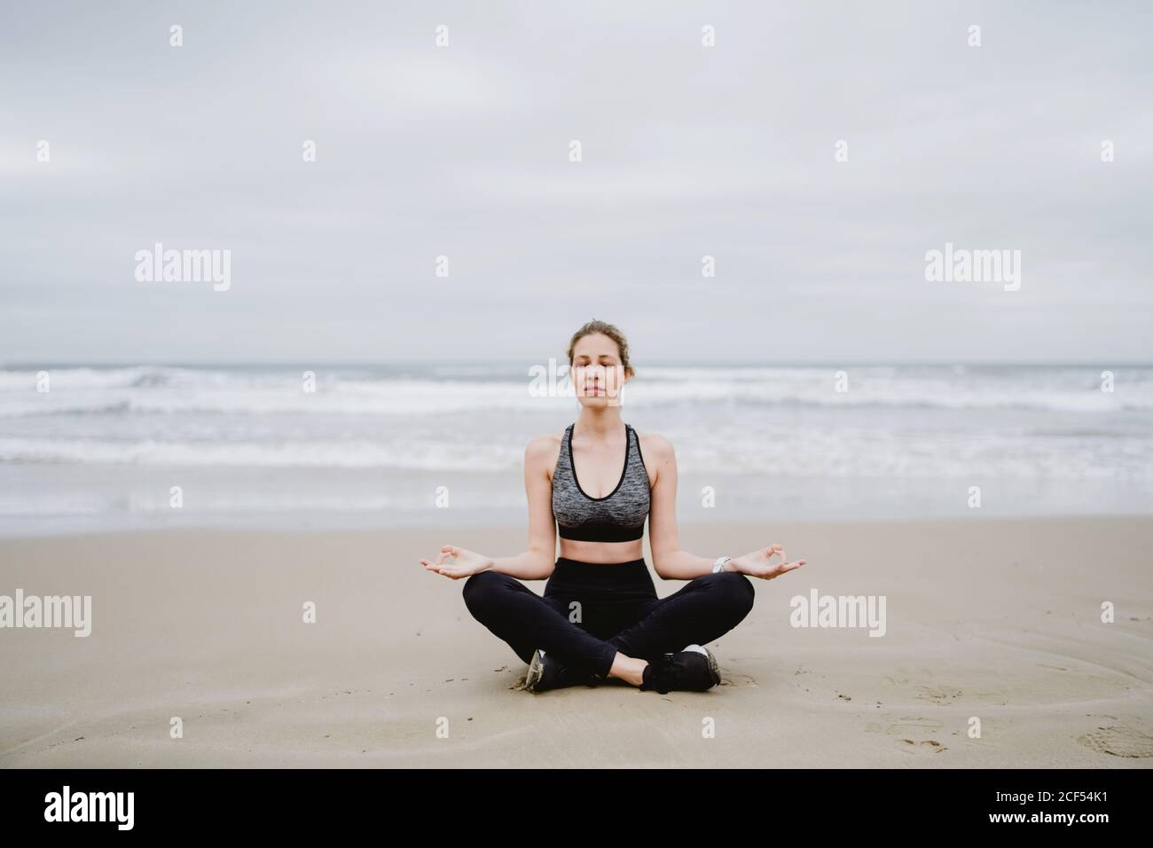 Young slim female in black top and leggings sitting on lotus position at the beach with closed eyes while meditating Stock Photo