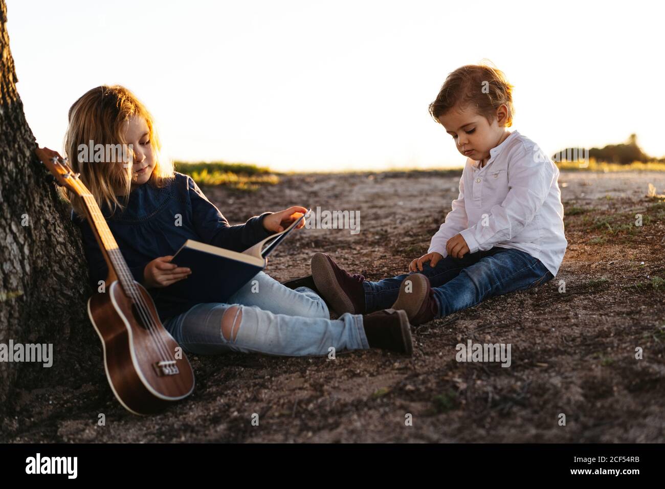 Side view of adorable little girl reading interesting story to younger brother while sitting together under tree with ukulele guitar in summer day in countryside Stock Photo