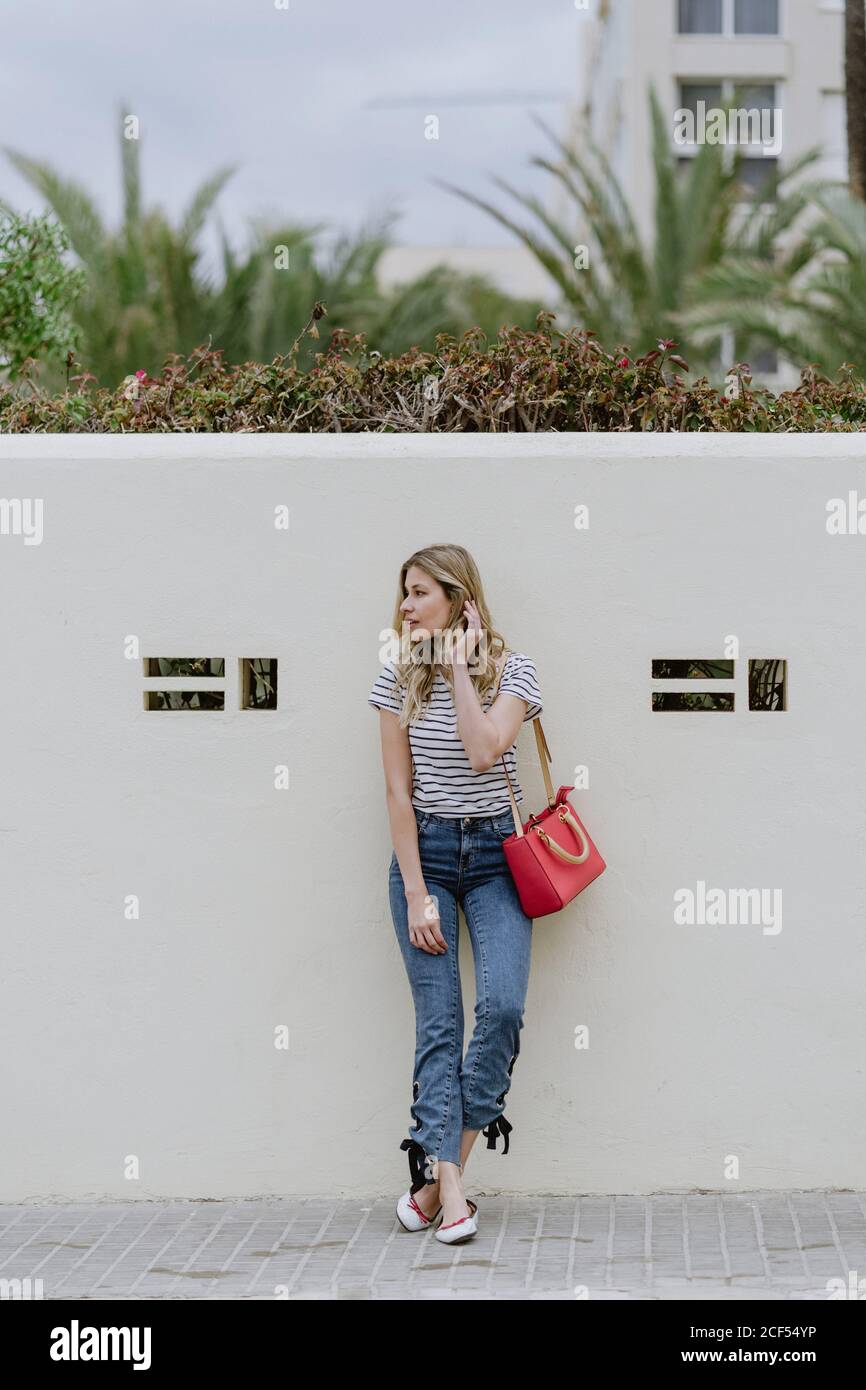 Charming young long haired blonde female with red shoulder bag wearing striped shirt and jeans standing and looking away Stock Photo