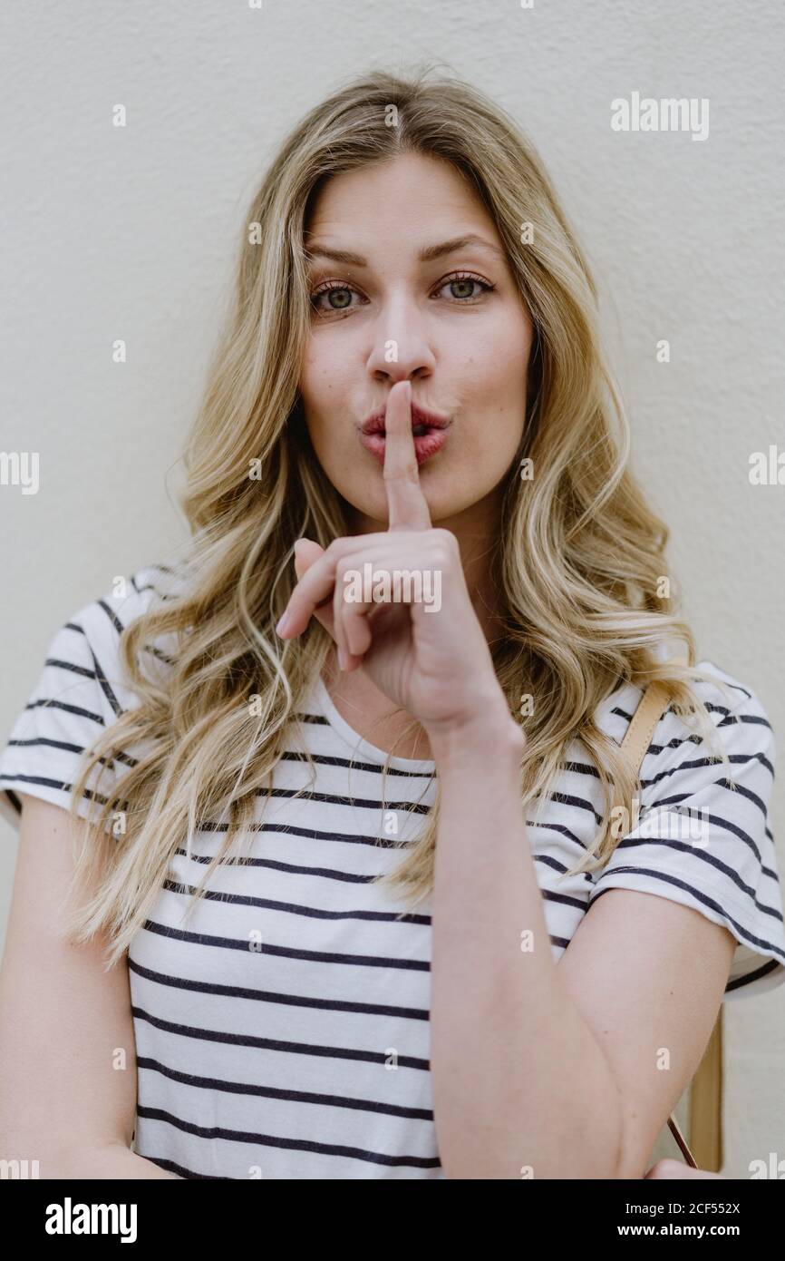 Attractive young female with long fair hair wearing casual striped shirt showing silence sign with finger on lips Stock Photo