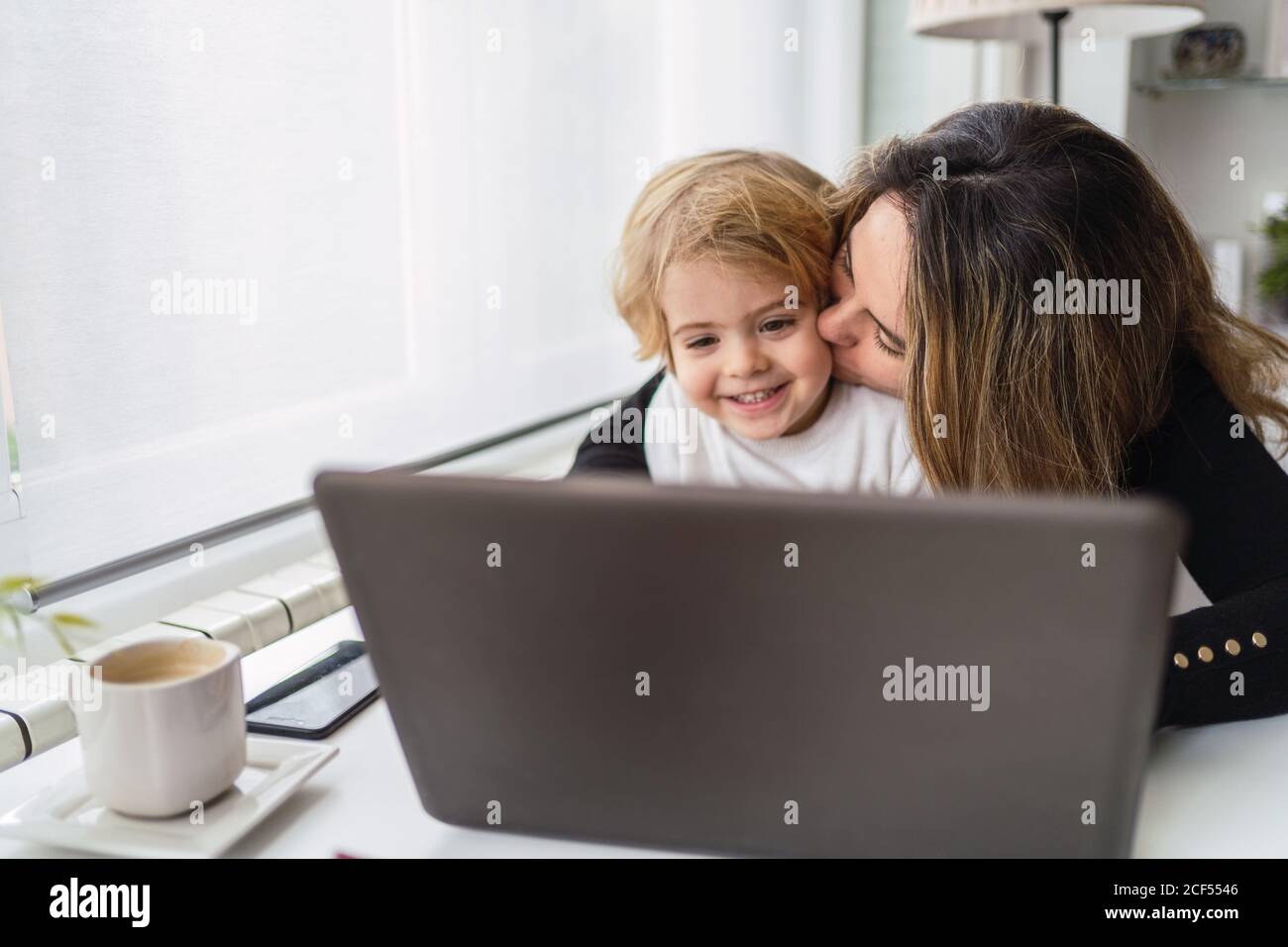 Female remote employee holding curious little child on knees while sitting at table and working with laptop at home Stock Photo