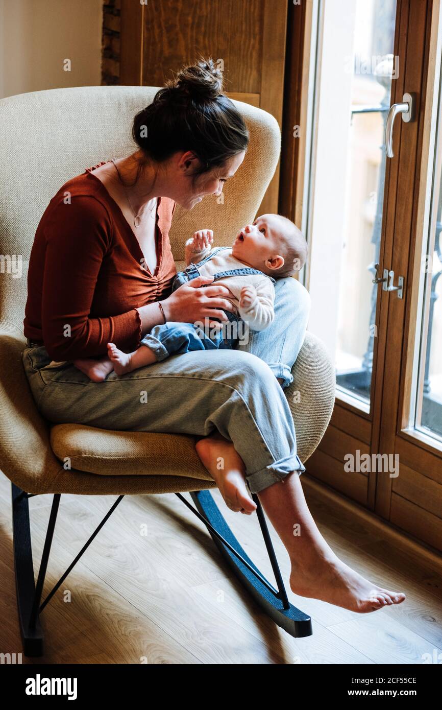 Full body barefoot Woman in casual clothes smiling and using baby talk with adorable infant while sitting on comfortable chair near window at home Stock Photo