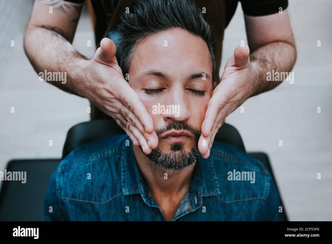 Crop barber doing face massage to handsome stylish male with closed eyes sitting in chair Stock