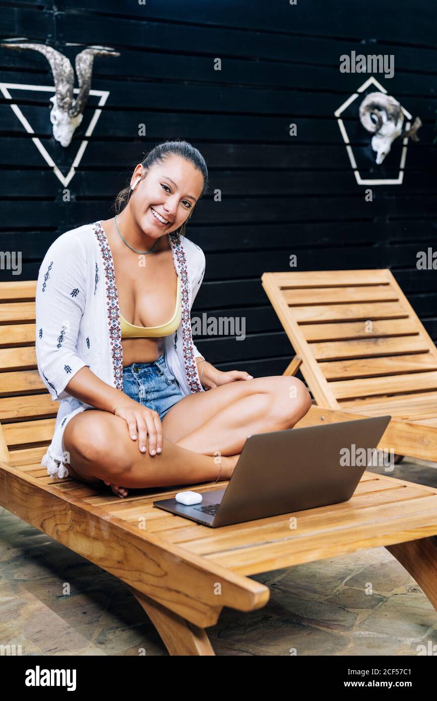Cheerful Woman in casual clothes browsing laptop while sitting with crossed legs on wooden deck chair at tropical terrace Stock Photo