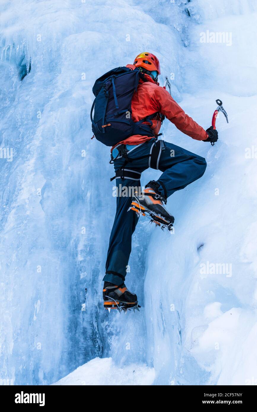 Back view of person in warm clothes with backpack using tools to climb frozen mountain glacier